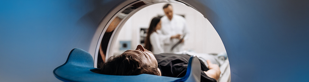 Pacient cu capul intr-un aparat RMN sau CT, cu medici discutand rezultatele in fundal.