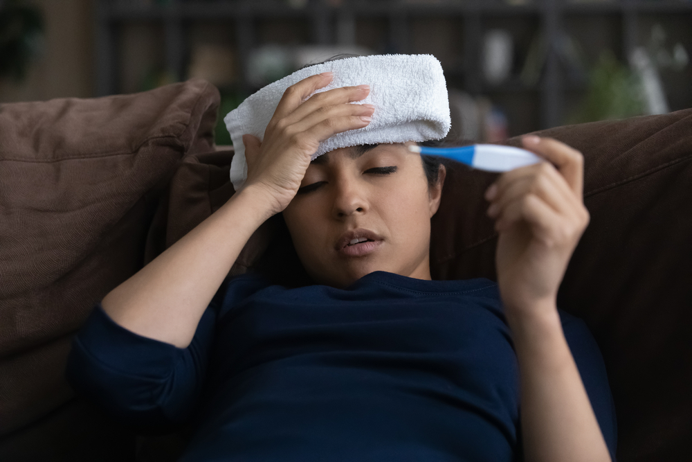 femeie tanara cu febra tine in mana un termometru si are o compresa aplicata pe frunte