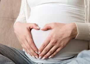 Burta unei femei insarcinate asezata pe o canapea, cu mainile pe burta formand o inimioara