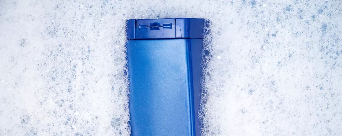 Recipient albastru de sampon plutind intr-o cada cu apa si spuma, simbolizand produsele de igiena personala.