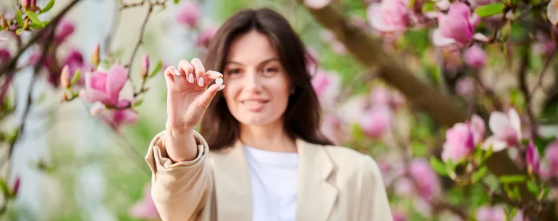 Femeie in aer liber care prezinta simtome de alergie sezoniera in timpul primaverii si care arata spre camera o pastila cu efect antihistaminic pentru