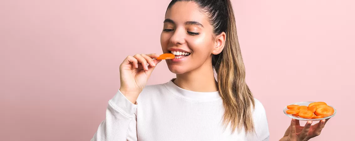 Femeie frumoasa zambind in timp ce mananca bucatele de morcovi dintr-un bol, ca sursa naturala de beta caroten