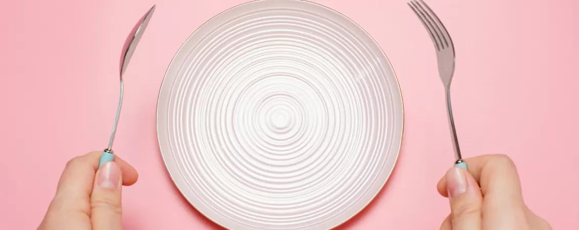 O femeie tinand in maini o lingura si o furculita langa o farfurie goala pe un fundal roz.