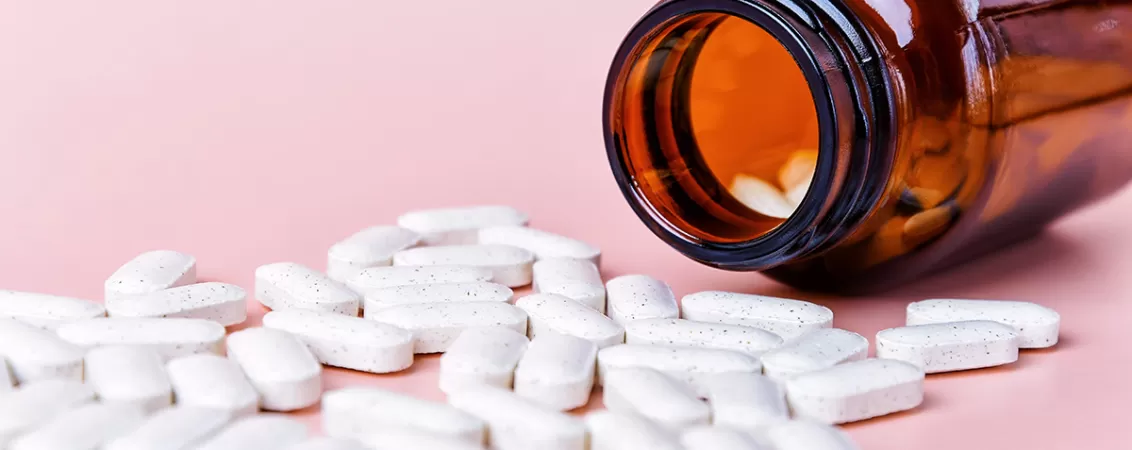 Sticla de chihlimbar rasturnata cu comprimate albe de vitamine imprastiate pe o suprafata roz