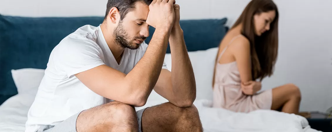 Bărbat trist, așezat pe pat, în timp ce partenera stă lângă el, ilustrând probleme sexuale în cuplu.