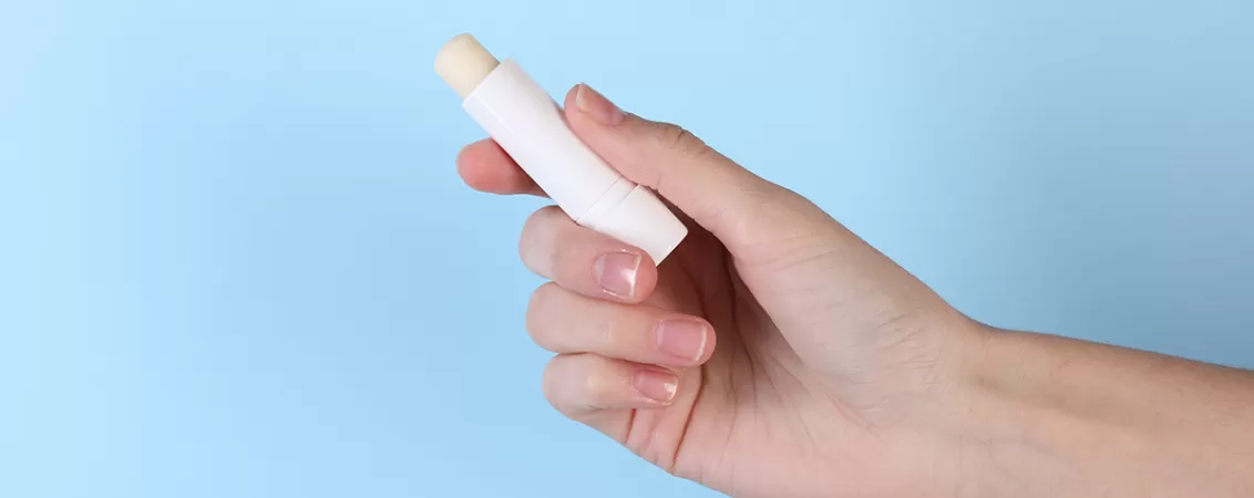 Femeie care tine in mana un balsam de buze pe un fundal albastru deschis, pentru hidratare si ingrijire