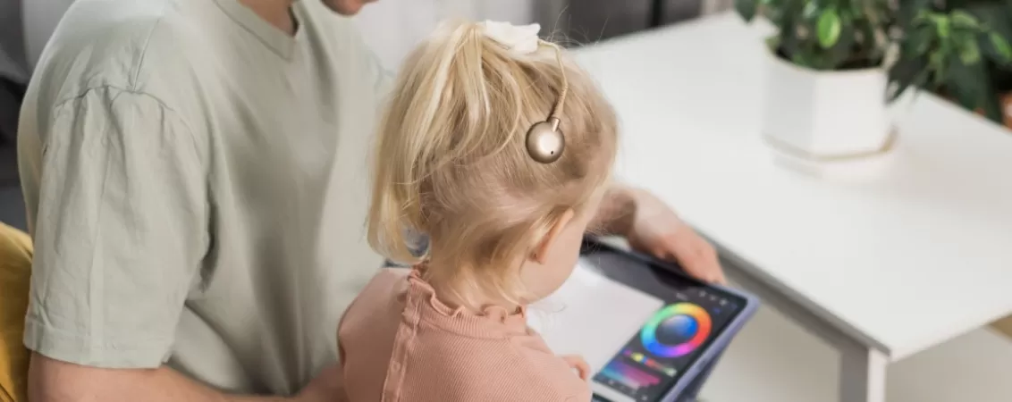 Prim-plan din spate cu o fetita care poarta un implant cohelar si care sta pe genunchiul tatalui, amandoi uitandu-se pe o tableta