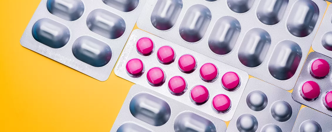 Vedere de sus a unor blistere cu pastile rotunde roz si unele ambalate cu gri, asezate pe un fundal galben