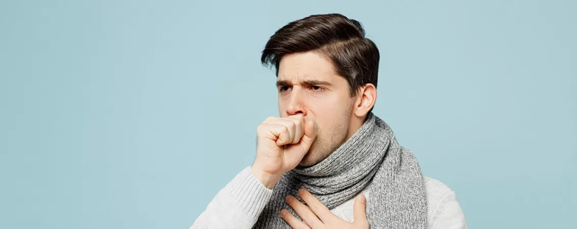 Tanar bolnav imbracat in hanorac gri care sta acasa si tuseste ca simptom de raceala sau gripa