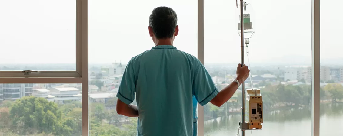 Pacient adult internat in spital, care tine in mana stativul cu perfuzie in timp ce se uita pe geam si primeste tratament intravenos