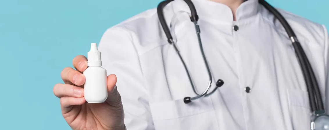 Picaturi medicale cu pilocarpina in mana unui medic pe fundal albastru, ilustrand administrarea precisa a picaturilor oftalmice pentru tratamente oftalmologice.