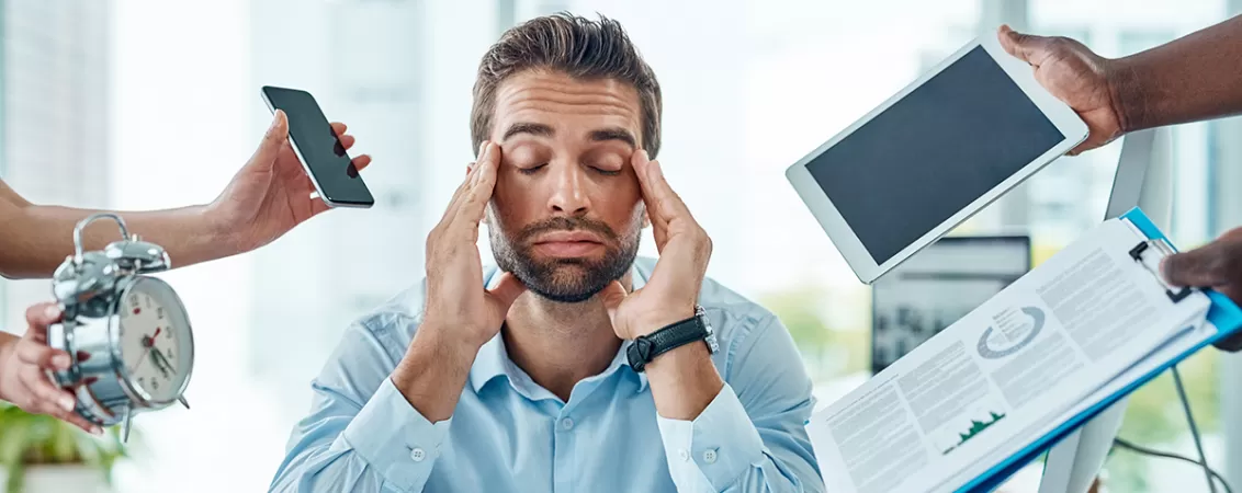Maini care intind laptop, telefon, ceas si doare spre un barbat care sta la birou cu mainile la tample, ilustrand suprasolicitarea profesionala.