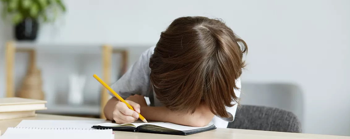 Baietel cu o tulburare de invatare cu capul pe birou, cu un creion in mana si un caiet gol in fata.