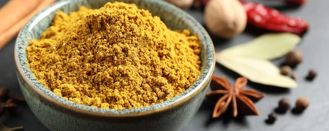 Pudra de curry intr-un bol, cu alte condimente in jur.