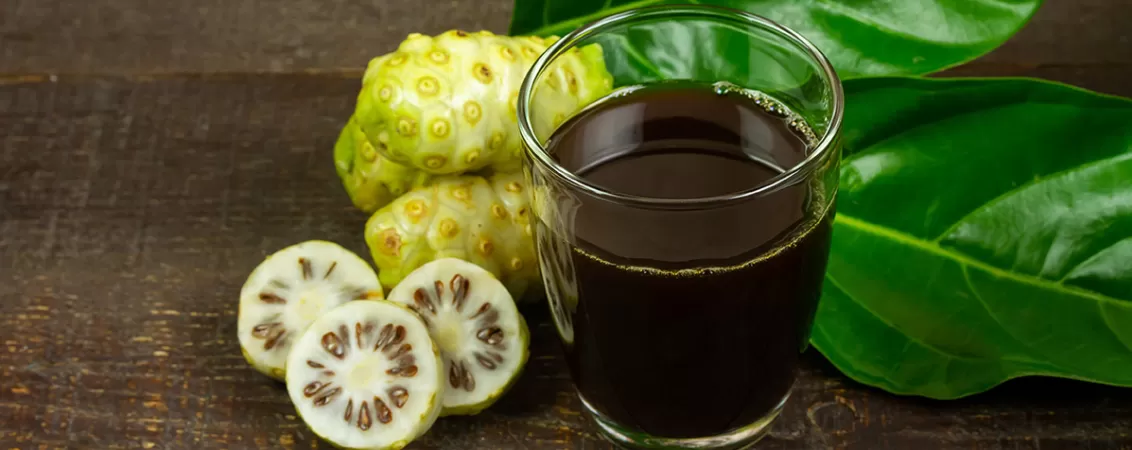 Suc de noni in pahar transparent, cu fructe noni langa.