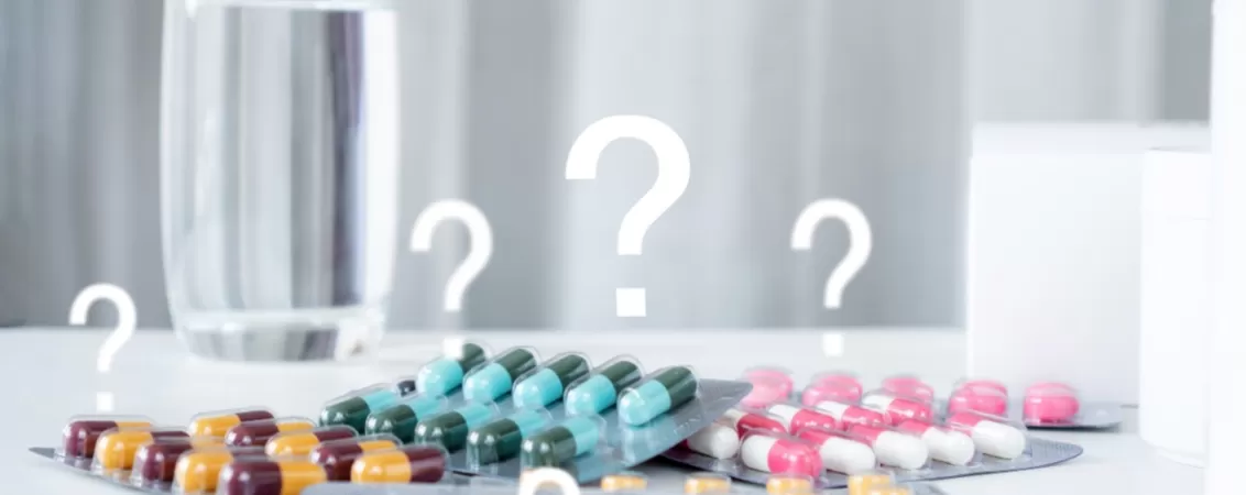 Diferite antibiotice pe o masa, langa un pahar cu apa si cu semne de intrebare deasupra
