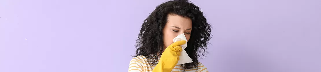 Femeie care poarta manusi si tine in mana o sticla cu detergent si se sterge la nas cu un servetel in urma unui stranut, reprezentand alergia la detergent