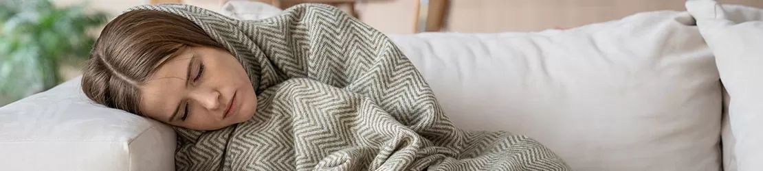 Femeie cu ochii inchisi stand pe canapea acasa, acoperita cu o patura pe cap, care prezinta simptome de sindrom serotoninergic