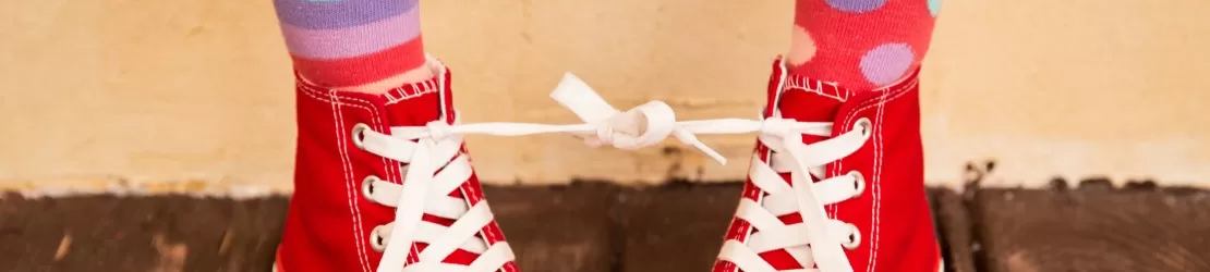 Picioarele unui copil incaltat cu bascheti rosii si sosete colorate, cu sireturile legate intre ele