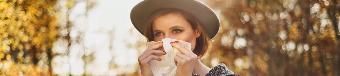 Femeie intr-un parc, toamna, care sufera de rinoree si isi sterge nasul cu un servetel