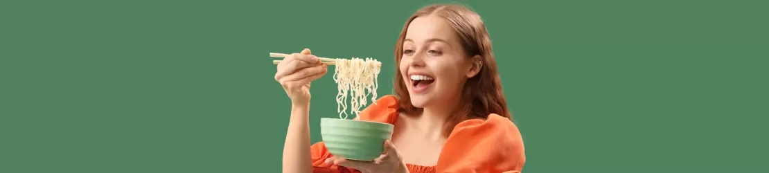 Femeie tanara fericita in timp ce mananca noodles dintr-un bol, ilustrand o masa rapida si obiceiuri alimentare din viata de zi cu zi.